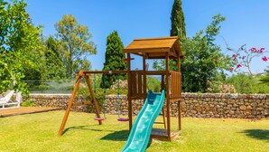 A wooden swing set with a slide in a backyard with a stone wall and greenery. - Country House 'Els Oscols 53' with Sea Views, Wi-Fi and Air Conditioning (Alcudia)
