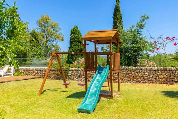 A wooden swing set with a slide in a backyard with a stone wall and greenery.