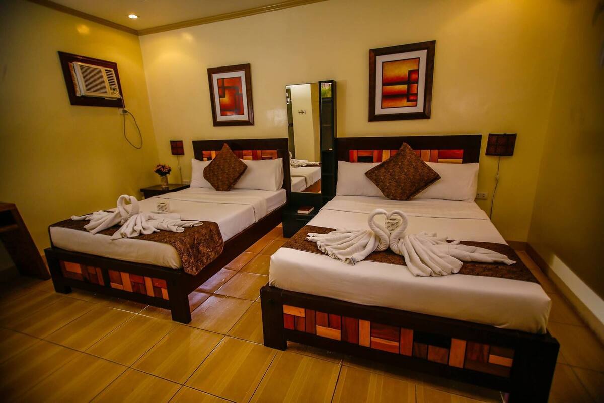 A hotel room with two beds, each with a wooden headboard, white linens, and towels arranged in a swan shape. There is a mirror on the wall between the beds, and artwork above each bed.