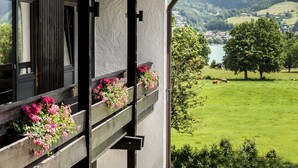 Un balcone con ringhiere in legno e fioriere piene di fiori rosa, con vista su un paesaggio verde con un lago e delle colline.