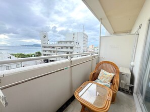 A balcony with a wicker chair and a small table, overlooking a cityscape and the sea.