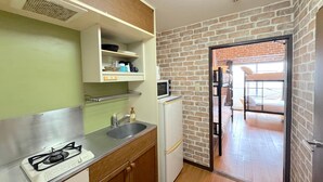 Une cuisine compacte équipée d’une cuisinière à gaz, d’un évier et d’un réfrigérateur blanc. On y trouve un mur en briques et une porte donnant sur un balcon.