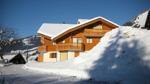 A wooden chalet with a snow-covered roof, a balcony, and a clear view of the surrounding snowy landscape.