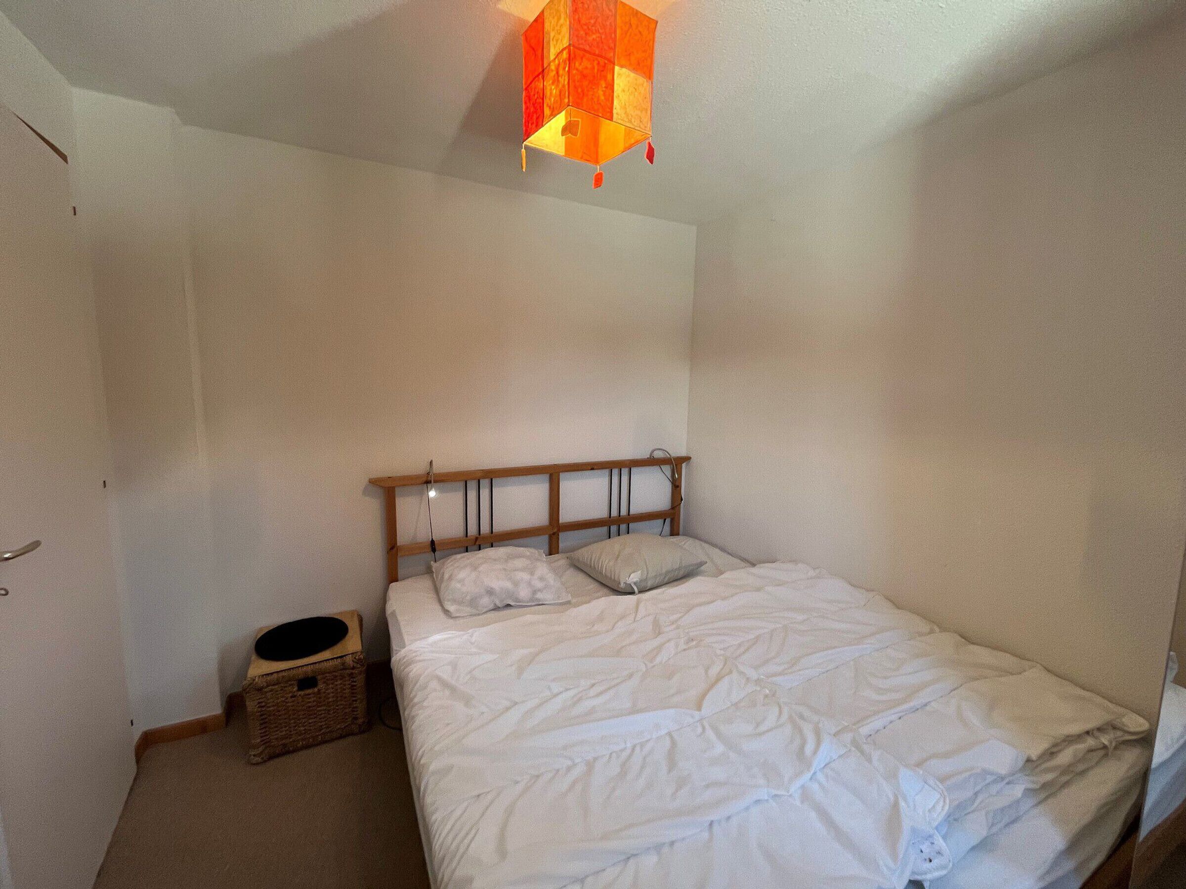 A bedroom with a wooden headboard, a single bed with white bedding, a small wicker basket, and a hanging light fixture.