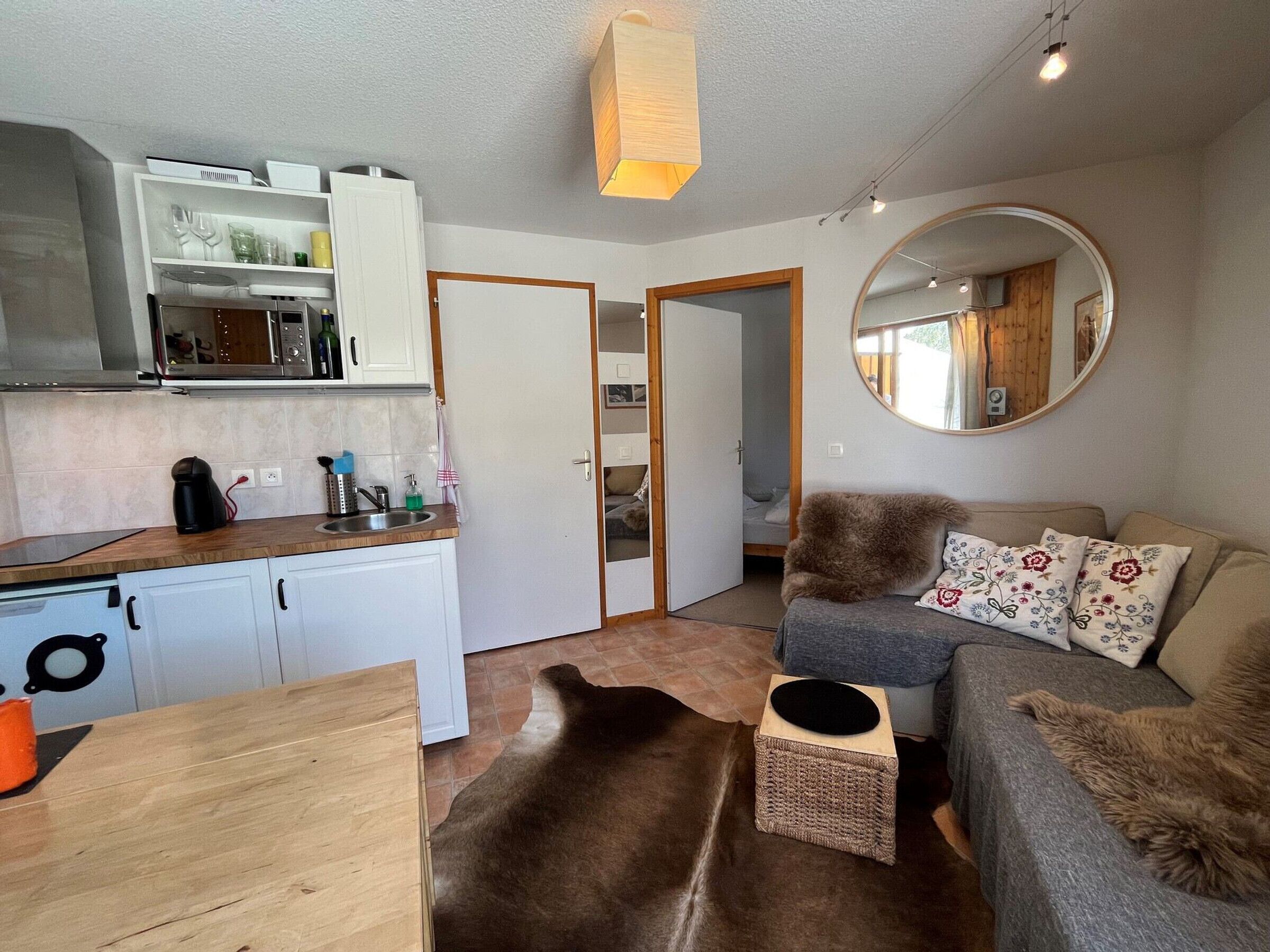 A compact living space with a kitchenette, a sofa with decorative pillows, a round mirror, and a small rug.