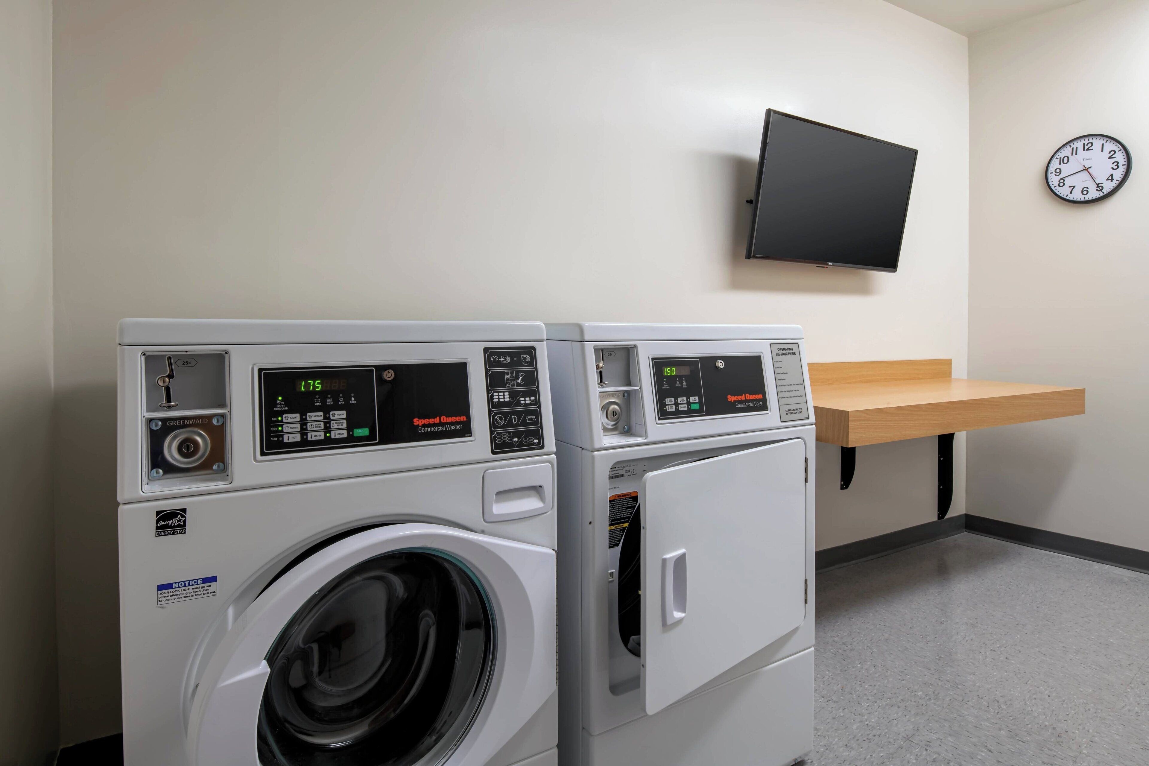 Deux machines à laver à chargement frontal dotées d’écrans numériques, un téléviseur fixé au mur et une horloge sur le mur.