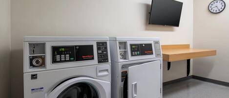Deux machines à laver à chargement frontal dotées d’écrans numériques, un téléviseur fixé au mur et une horloge sur le mur.