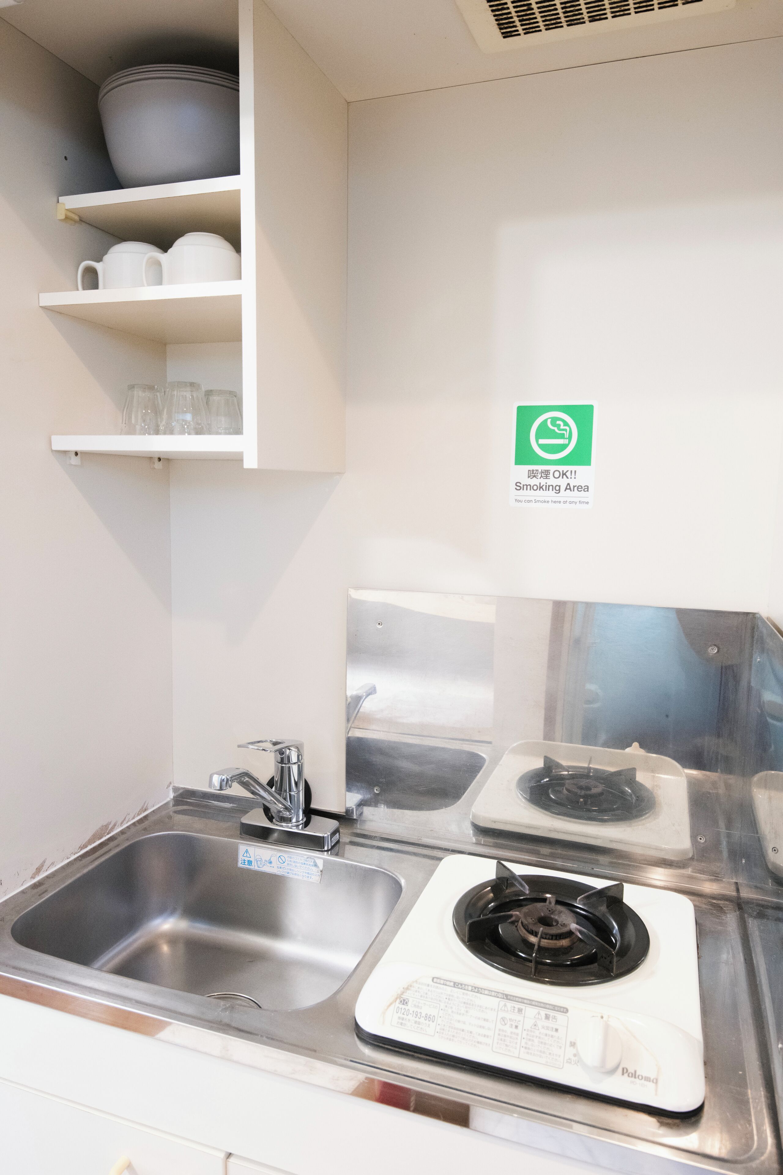 Une cuisine équipée d’un évier en acier inoxydable, d’une cuisinière et d’une étagère où sont rangées des tasses et des bols. Un panneau indique l’emplacement de la zone fumeurs.