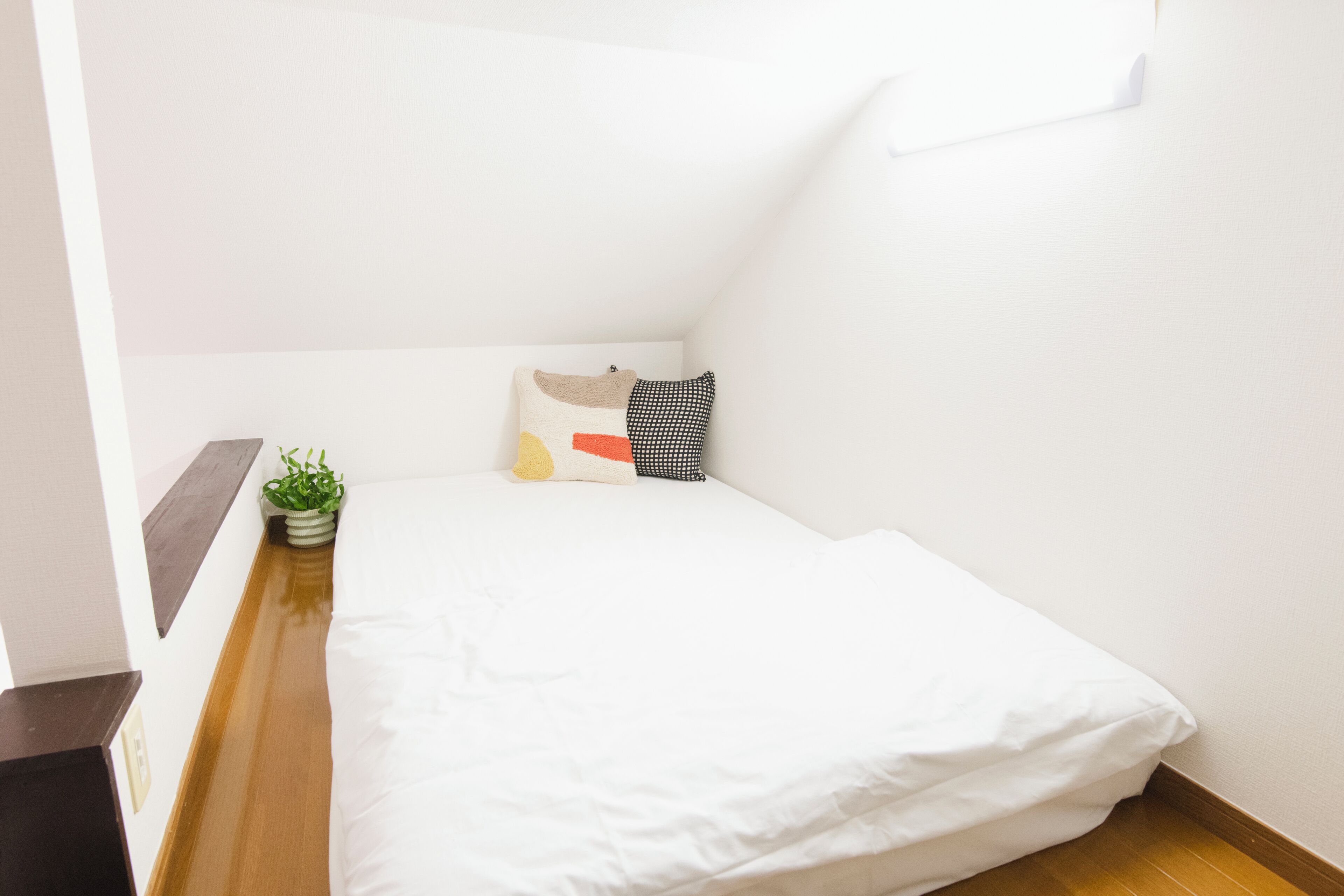 A small, neatly made bed with white bedding in a corner room with white walls and a wooden floor.