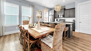 A dining area with a wooden table and wicker chairs, a kitchen with dark cabinets, and a white door. - ☀️Pool + Hot Tub-4 Bikes-Luxury Home on 30A-Sandy Cheeks (Santa Rosa Beach)