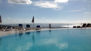 A swimming pool with lounge chairs and umbrellas, a metal railing, and a view of the sea.