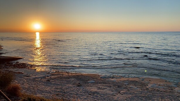 A beach with the sun setting over the water, rocky shoreline, and calm sea.