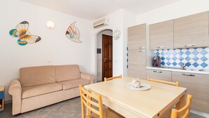 A living room with a beige sofa, a wooden dining table, and chairs. There is a kitchen area with wooden cabinets and a tiled backsplash.