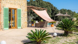 A stone building with green shutters, outdoor seating, a shaded area, and a pool.