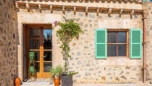 A stone building with a wooden door and green shutters, potted plants, and a tiled roof.