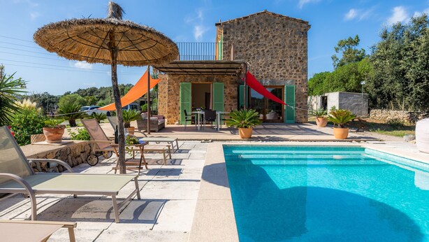 A poolside area with lounge chairs, a thatched umbrella, and a stone building with green shutters.