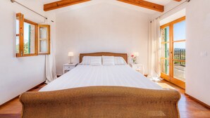 A bedroom with a large bed, wooden beams, a nightstand, a lamp, and a view through a glass door.
