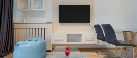 A modern living room with a glass coffee table, a blue bean bag, two black chairs, a flat-screen TV, and a white entertainment unit.