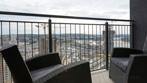 Un balcon avec deux fauteuils en osier, une vue sur un paysage urbain avec des bâtiments et une rivière, et une grue de construction visible au loin.