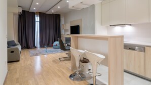 Un salon moderne avec un canapé, un coin repas comprenant une table et des chaises, une cuisine équipée de placards et d’un lave-vaisselle, et une grande fenêtre agrémentée de rideaux.