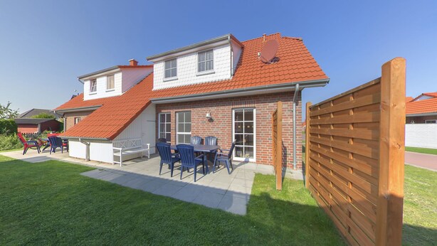 Une maison à deux étages avec un toit recouvert de tuiles rouges, une façade en briques et une cour arrière clôturée, aménagée avec du mobilier d’extérieur.