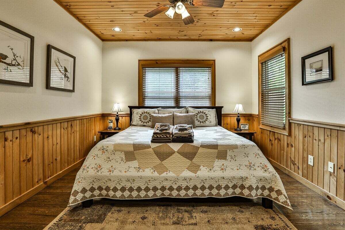 Een slaapkamer met een houten plafond, een groot bed met quilt, twee nachtkastjes en een raam met zonwering.