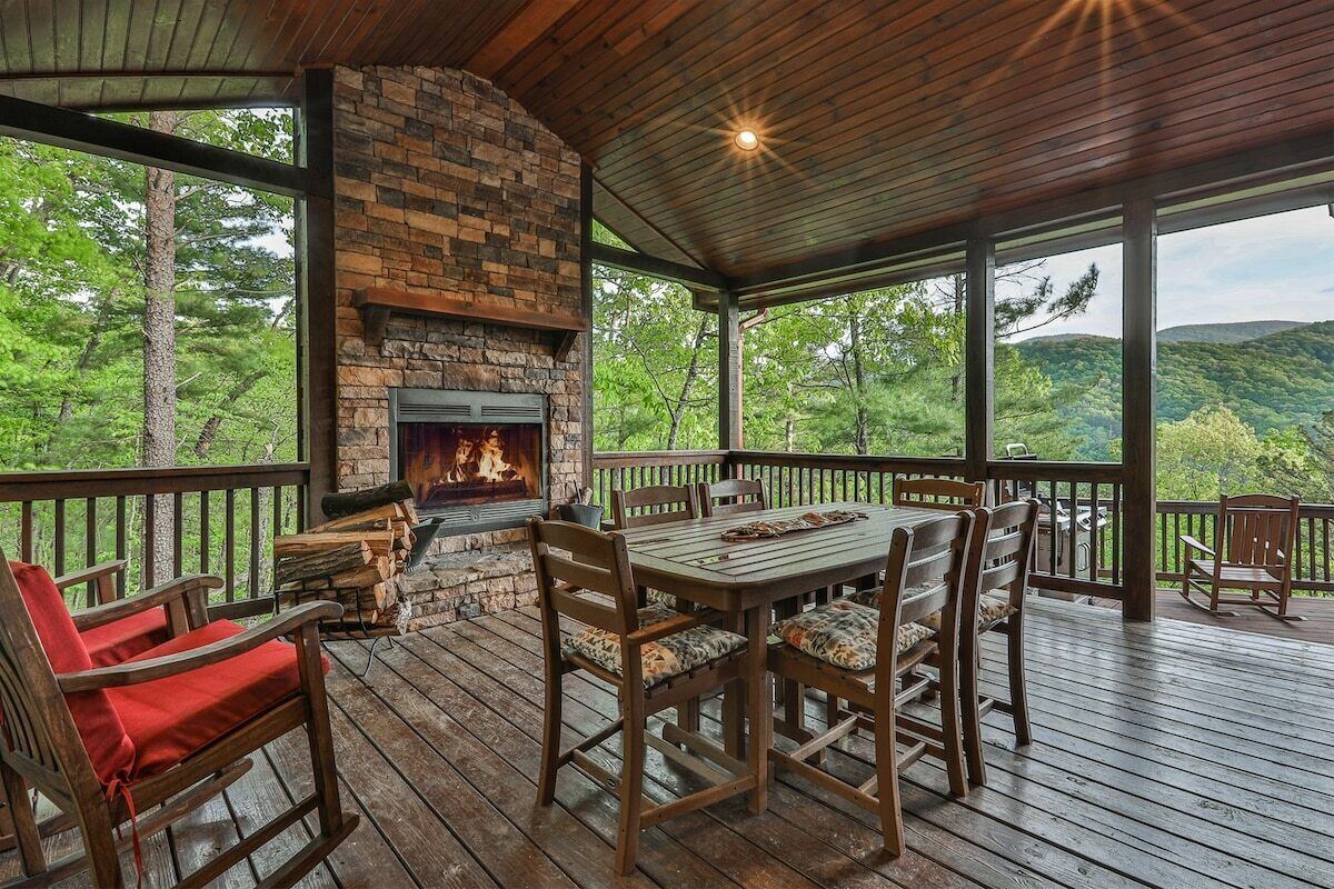 A covered outdoor area with a stone fireplace, wooden furniture, and a view of the surrounding forest.