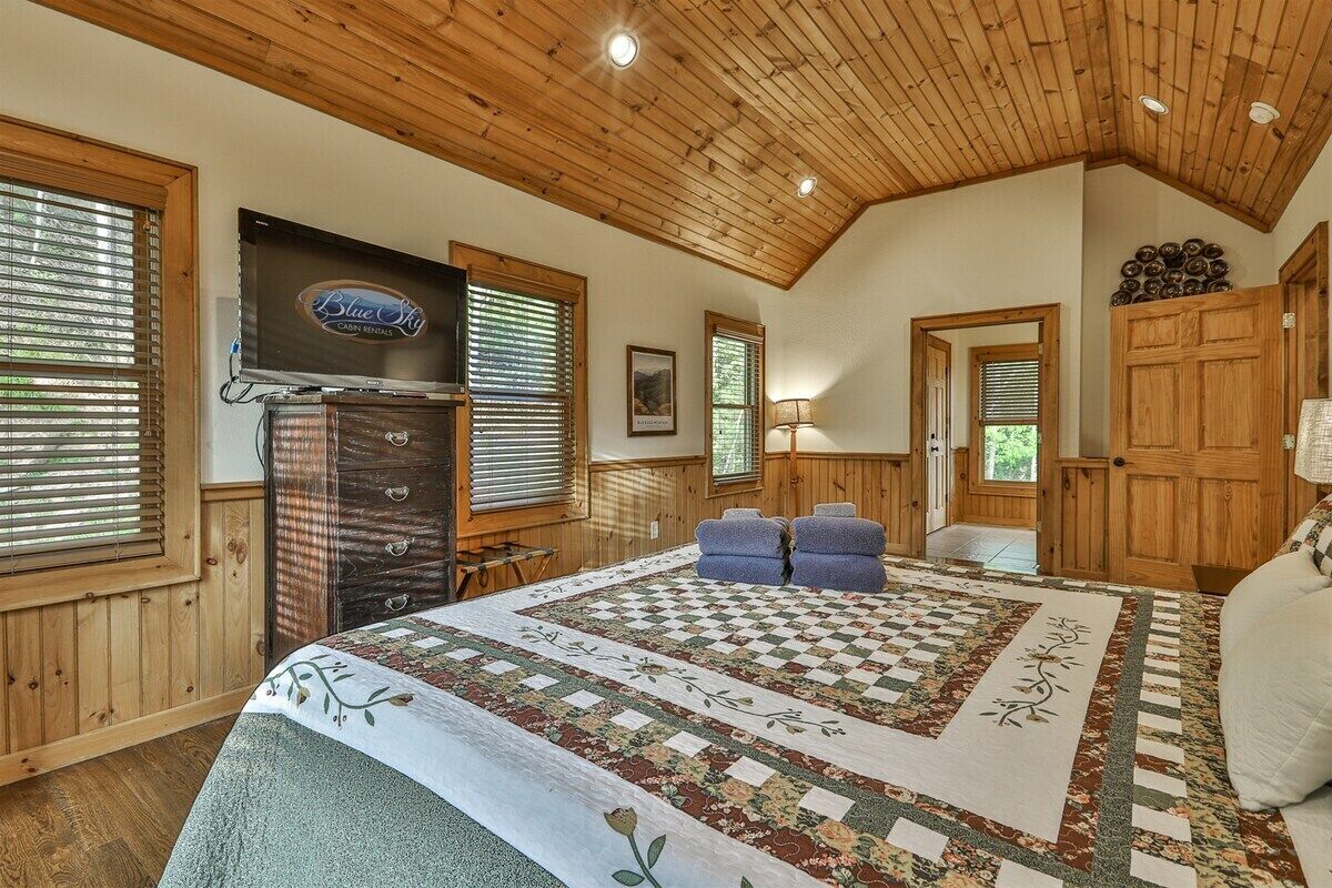 A cabin with a wooden ceiling, a fireplace, a television, a bed with a quilt, and a door with a wooden panel.
