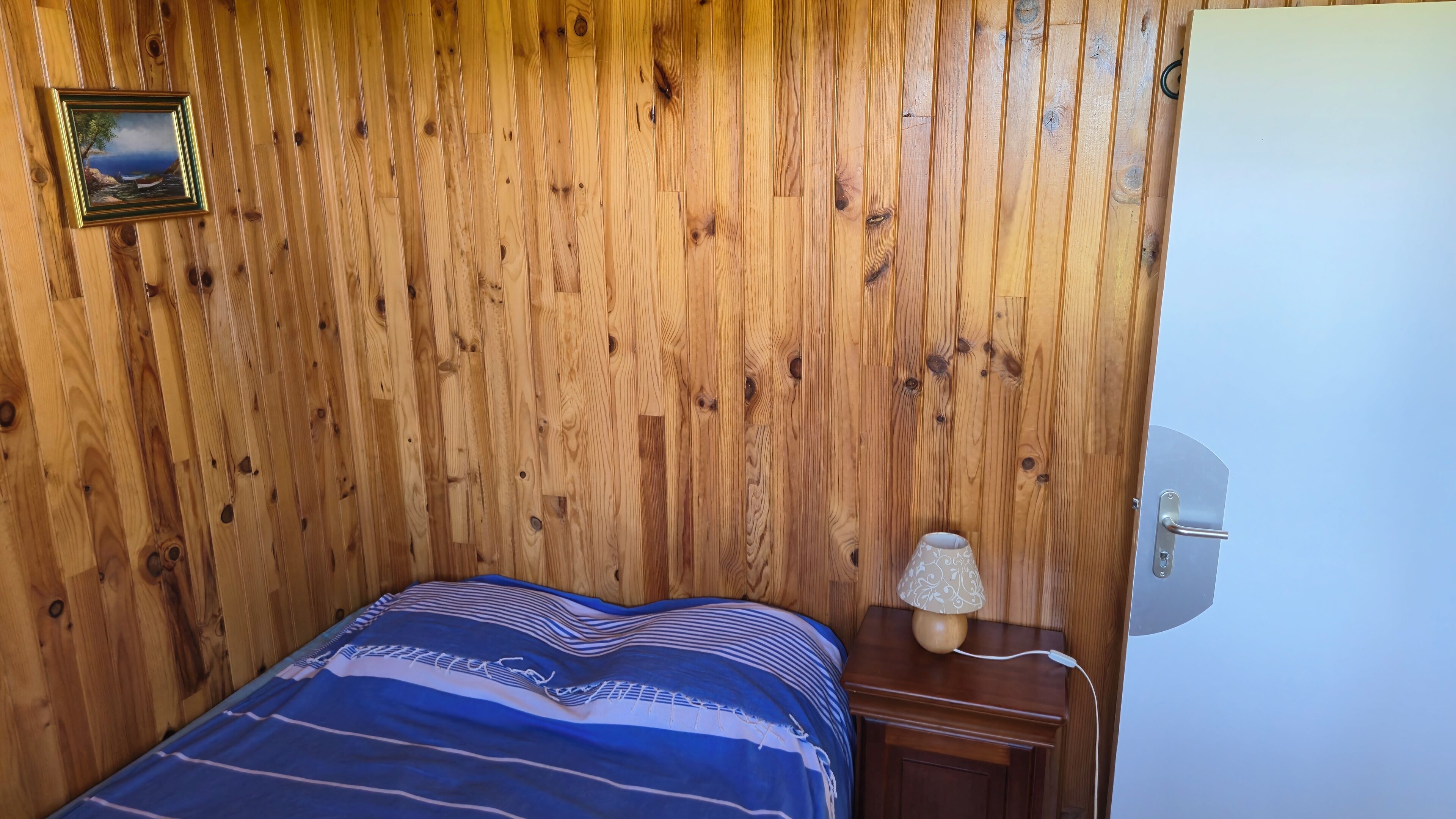 Habitació amb panells de fusta, un llit, una taula de nit, una làmpada i una porta.