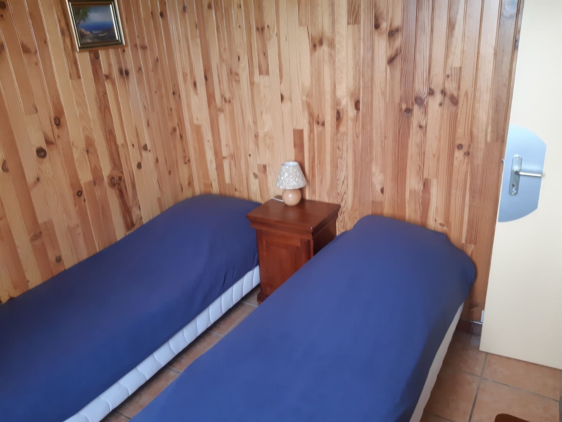 Habitació amb dos llits, panells de fusta a les parets, una petita taula de nit de fusta amb una làmpada i una porta amb tirador metàl·lic.