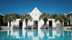 Zona de piscina con una piscina azul cristalina, palmeras y un edificio con un reloj y detalles decorativos.