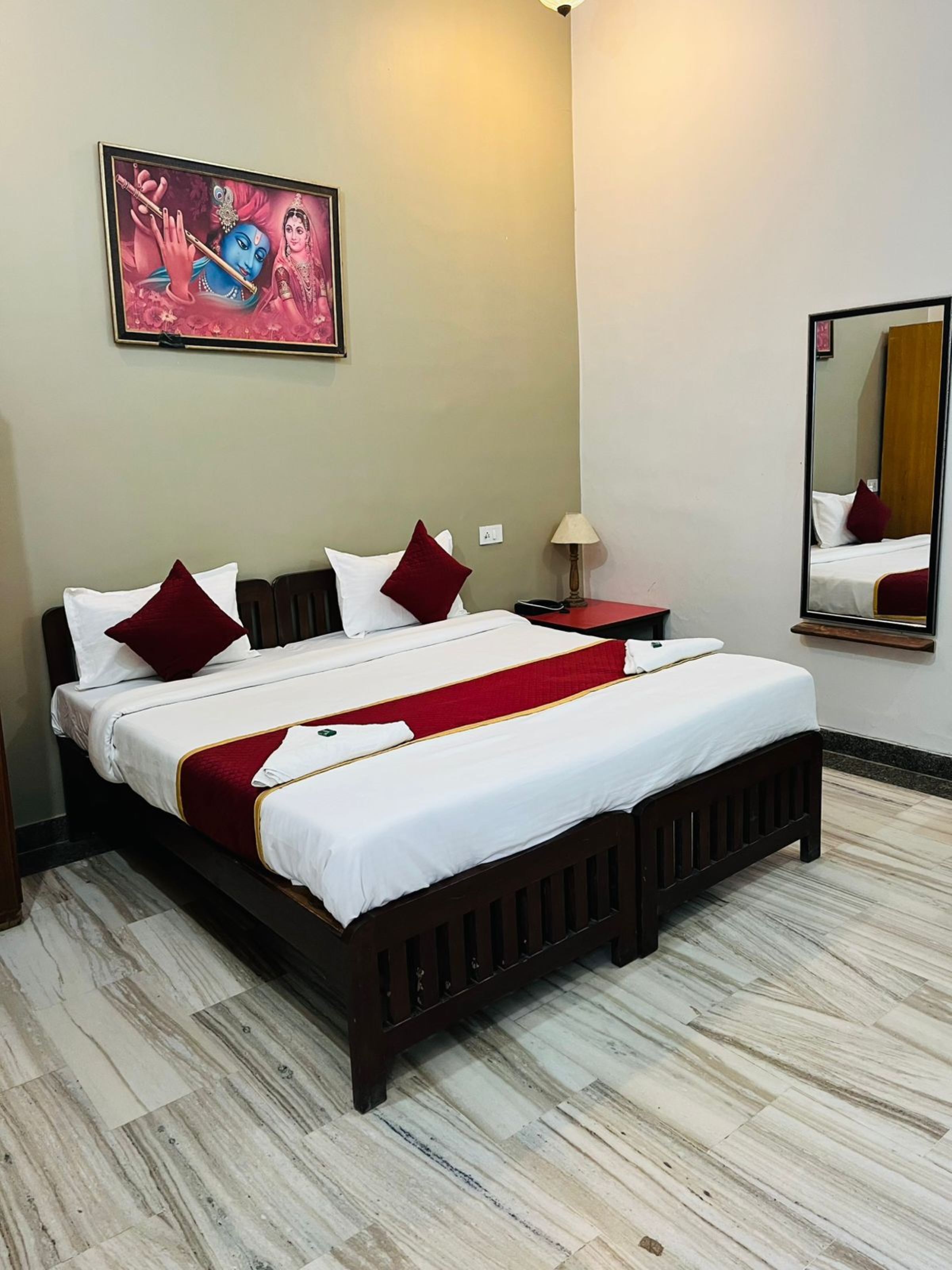 Habitación de hotel con cama doble, ropa de cama roja y blanca, cabecera de madera, mesita de noche con lámpara, espejo y un cuadro en la pared.