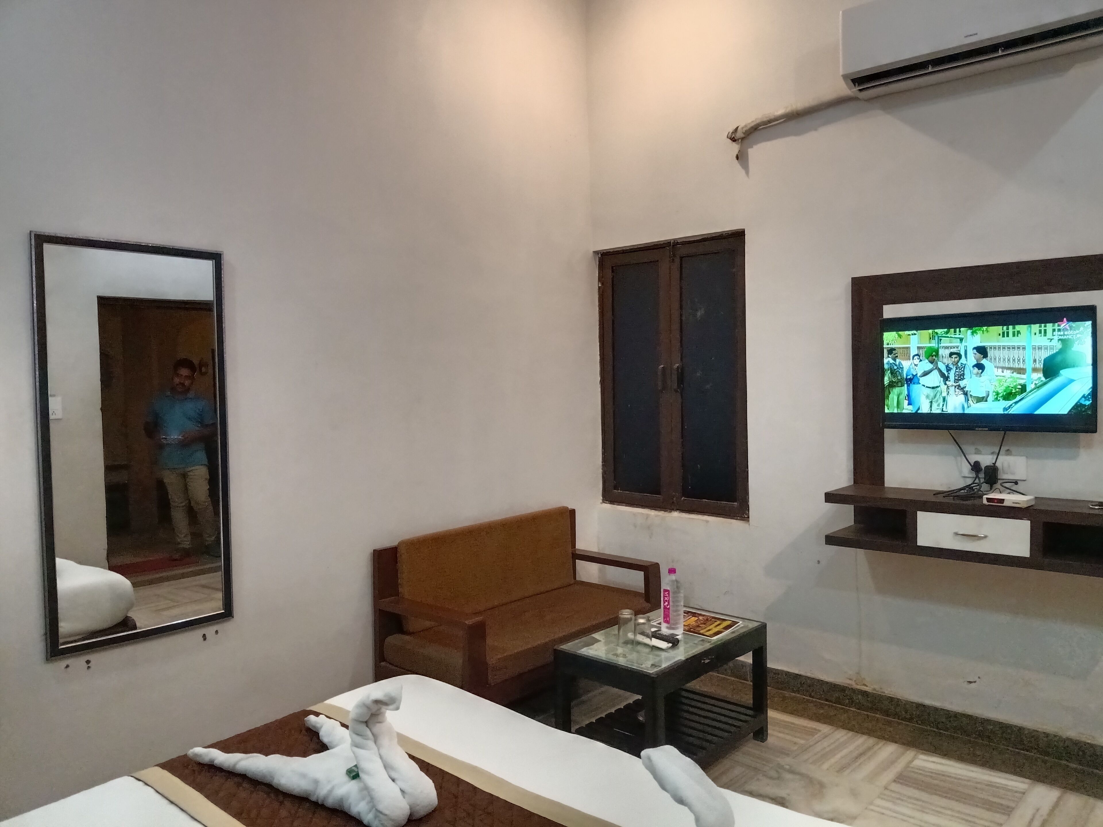 A hotel room with a bed, a TV, a mirror, a brown sofa, a small table with a bottle, and a window with a view.