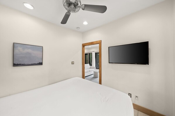Un dormitorio con cama, ventilador de techo, televisor montado en la pared y un cuadro enmarcado.