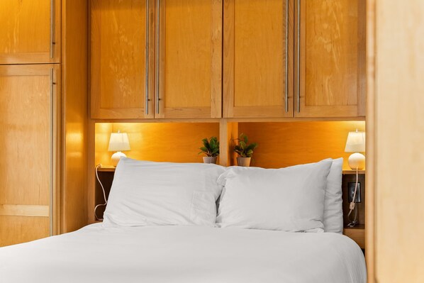 Una cama bien hecha con sábanas blancas, cabecera de madera, lámparas de noche y dos pequeñas macetas.