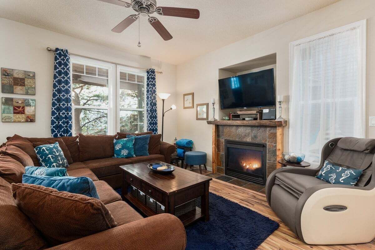 Een woonkamer met een bank, fauteuil, televisie, open haard en plafondventilator.