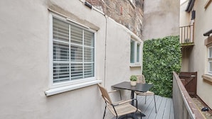 En balkon med et bord, stole, et vindue og en grøn hæk.