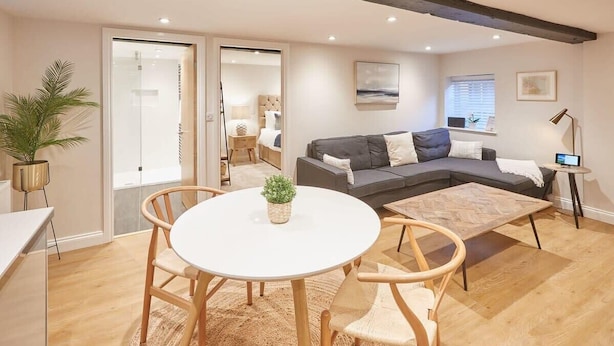 En moderne stue med en grå hjørnesofa, et rundt hvidt bord og træstole. Der er en potteplante på bordet, og rummet har trægulv og et stort vindue med persienner.