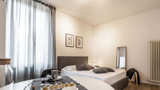 A hotel room with a dining table set for breakfast, a bed with a headboard, a bedside table, a mirror, and a radiator. - Emilia Suite Essence (Modena)