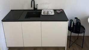 A compact kitchenette with a black countertop, a sink, and a small dining area with a chair. - Emilia Suite Essence (Modena)