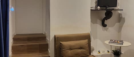 A living room with a sofa, a television on a shelf, a small table with a plant, and a staircase.