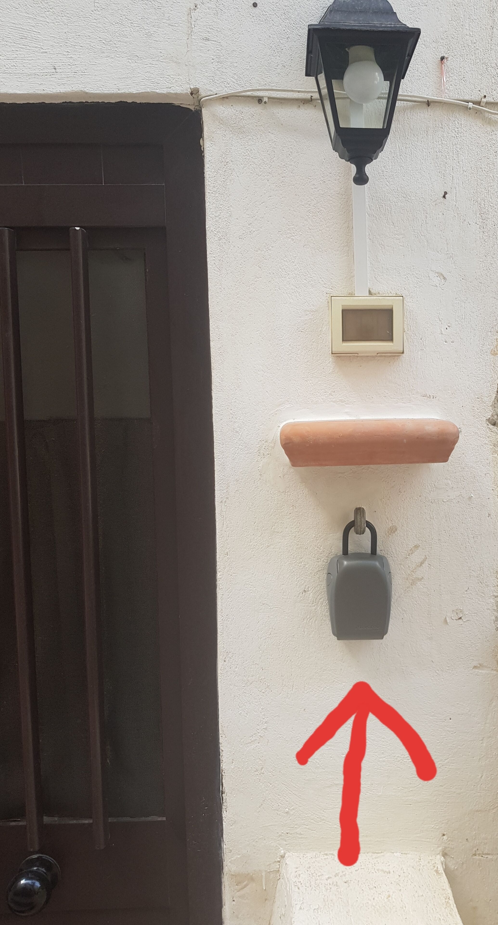 Uma porta com moldura escura, luminária fixada na parede, pequeno painel de controle e um cadeado.