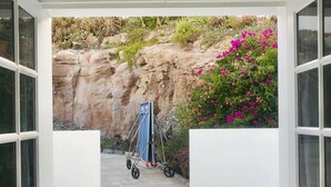 Abertura de porta que dá para um pátio com parede de pedra, uma cadeira dobrável azul e flores cor-de-rosa.