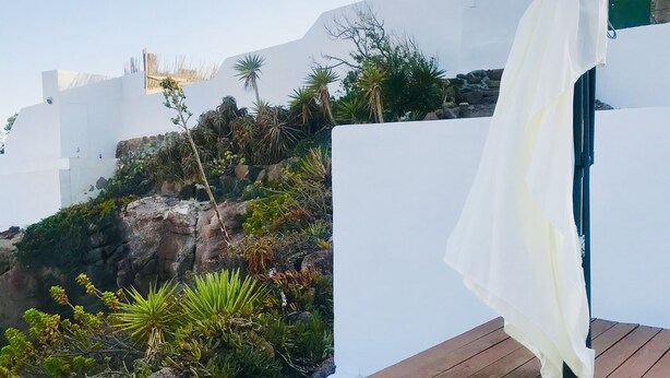 Mesa de madeira com um guarda-sol branco num terraço com paredes brancas e vista para uma paisagem rochosa com cactos e céu claro.