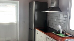 Una cocina con un refrigerador de acero inoxidable, armarios blancos, una encimera de madera y una ventana con persianas.