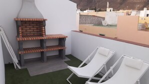 Un balcón con una parrilla de ladrillo, dos sillones blancos y vista a un paisaje urbano con edificios y montañas al fondo.