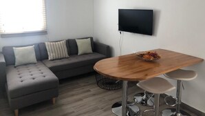 Una sala de estar moderna con un sofá seccional gris, una mesa de comedor redonda de madera con taburetes, un televisor de pantalla plana montado en la pared y una ventana con persianas.