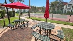 Un espacio al aire libre con mesas y sillas de metal verde, un paraguas rojo y una cancha de tenis al fondo.