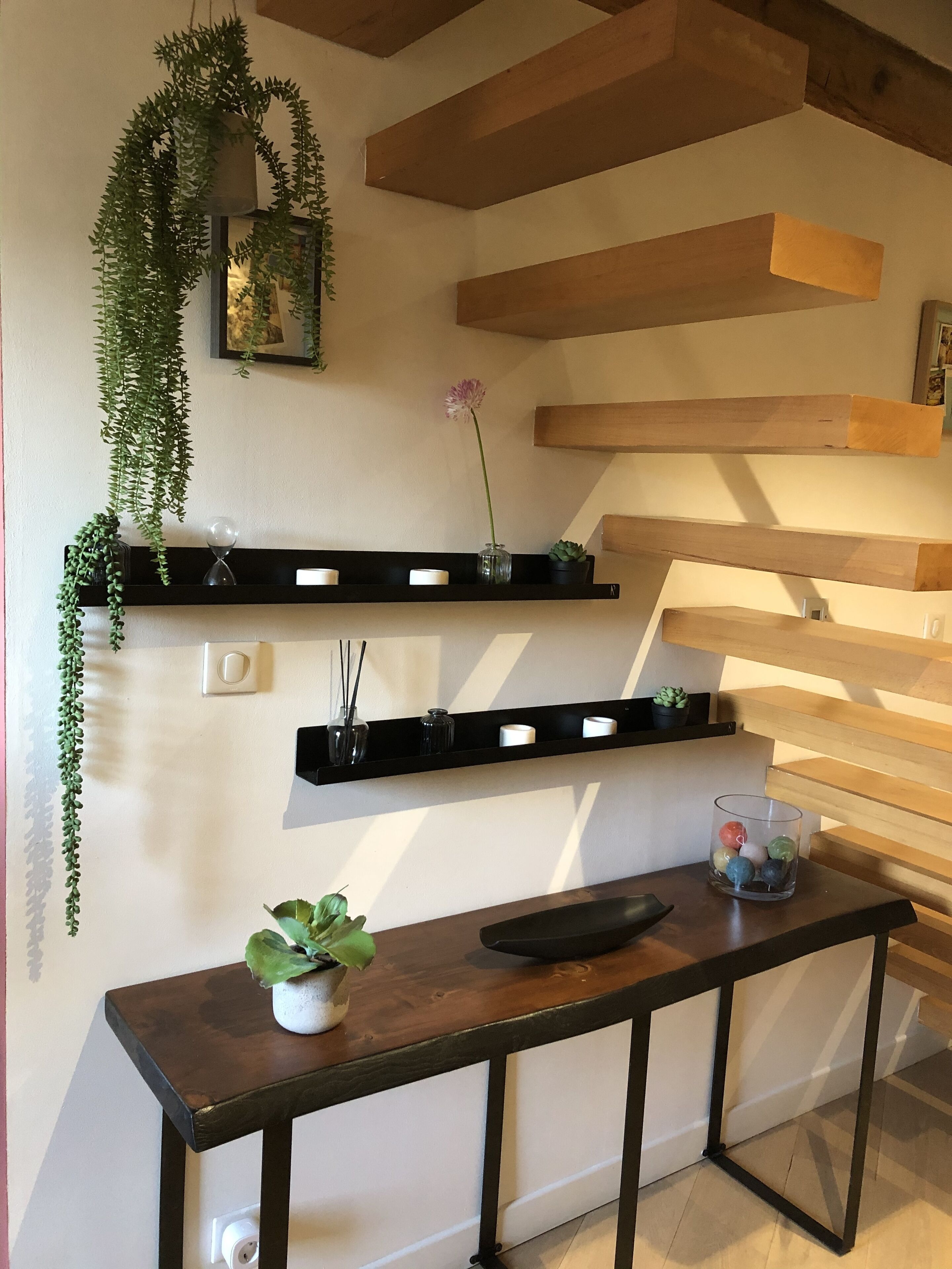 A modern interior with wooden beams, black floating shelves with plants and candles, a small table with a potted plant, and a glass container with colorful balls.