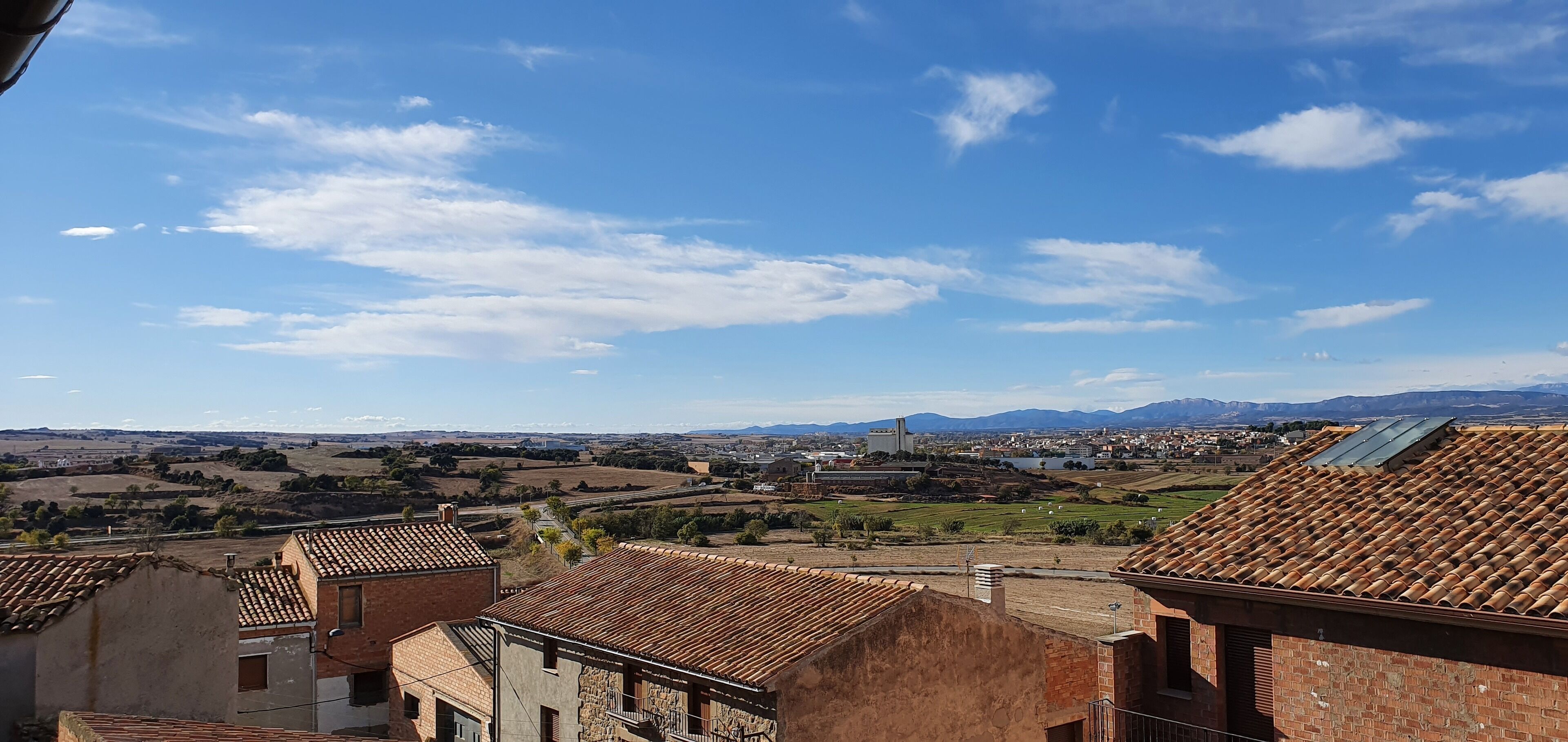 Paisaje rural con edificios de tejados terracota, una cadena montañosa distante y un cielo azul despejado.